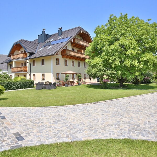 Exterior view of the farmhouse with a paved driveway, lawn, and outdoor terrace with seating, with a church in the background.