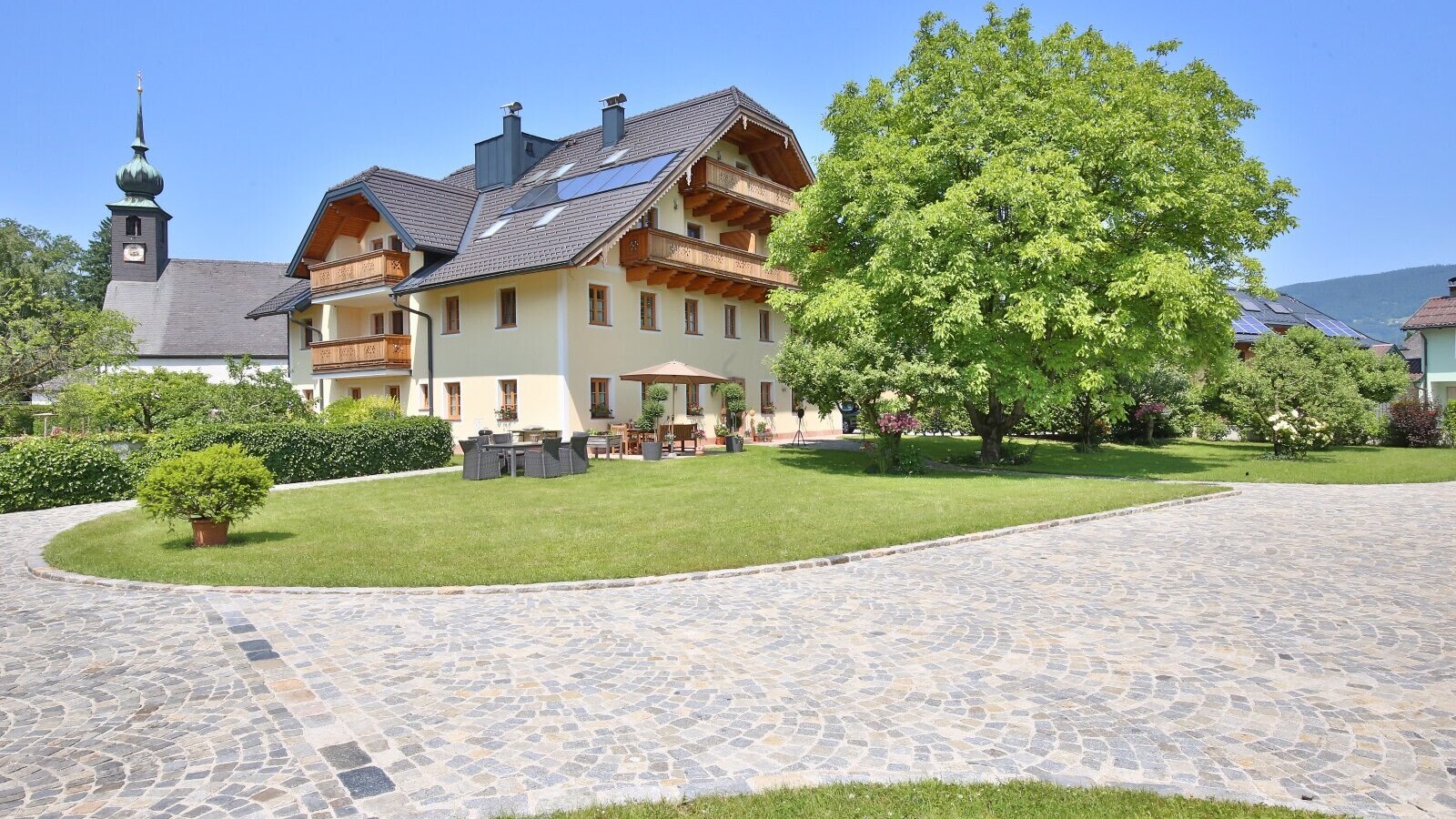 Exterior view of the farmhouse with a paved driveway, lawn, and outdoor terrace with seating, with a church in the background.