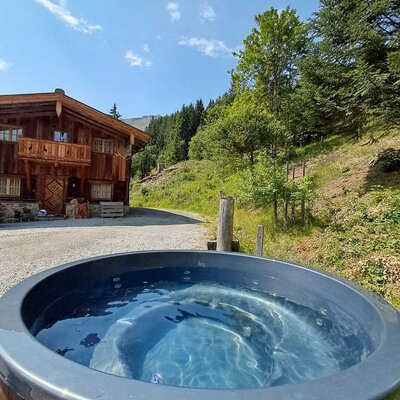 An inviting outdoor hot tub in front of the rustic Alm, surrounded by lush green forests.