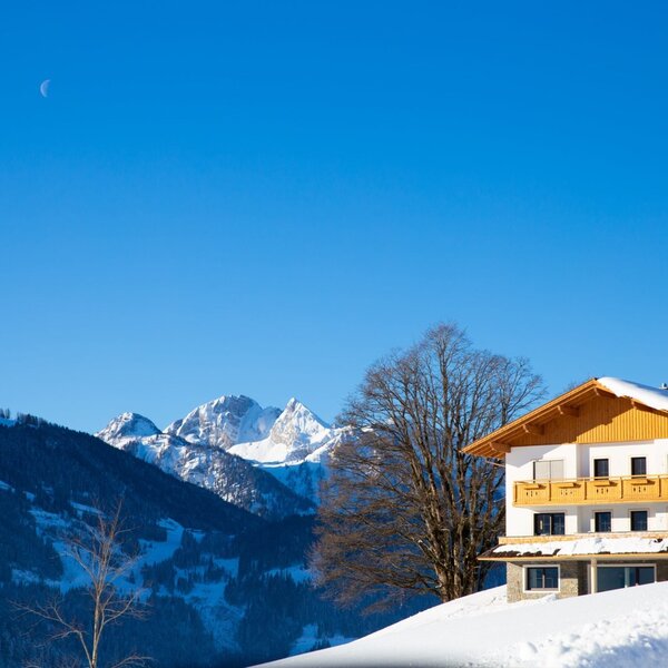 The farmhouse exterior in winter, featuring snow on the roof and balconies, with the snowy Salzburg mountains visible in the background.