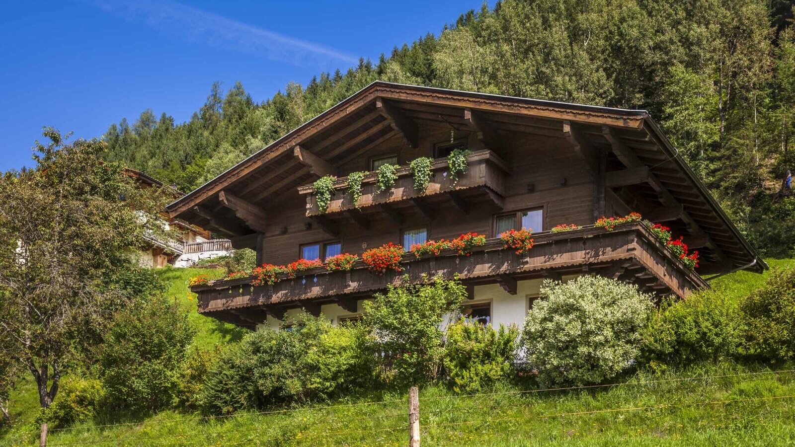 The farm house features a traditional wooden exterior with balconies decorated with red flowers and greenery, situated on a green hillside.