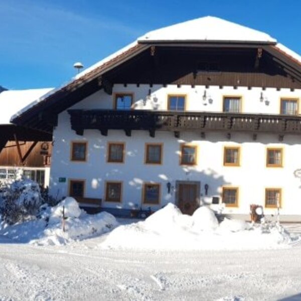 The snow-covered Farm House, featuring a prominent balcony, amidst a winter landscape with trees and mountains.