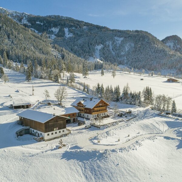 The farmhouse, a multi-story building with balconies, surrounded by snow-covered fields and mountains, with additional farm buildings nearby.