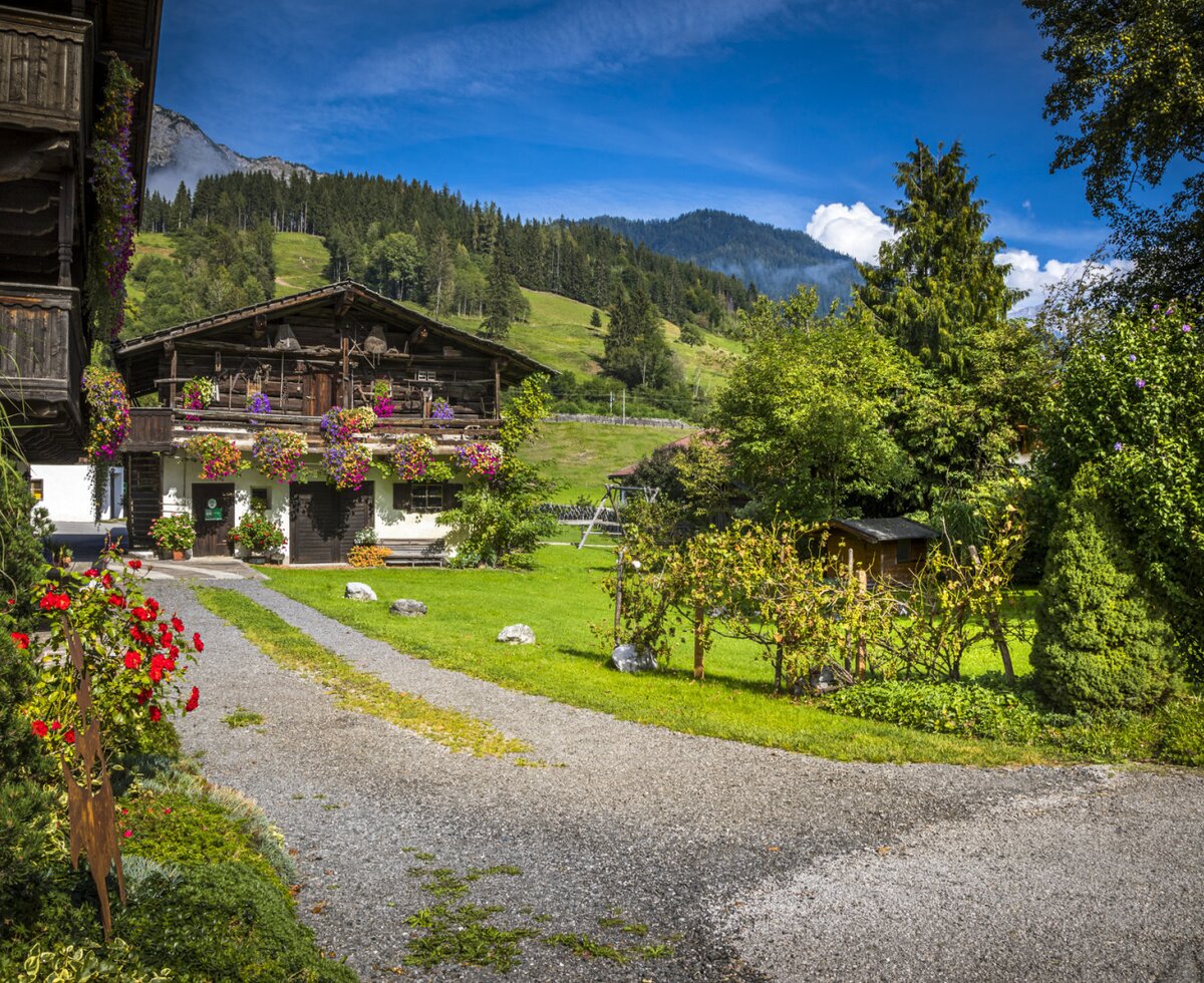 The farm house exterior, featuring flower-decorated balconies, a gravel driveway, a lawn, and a garden with a swing set, set against a mountain landscape.