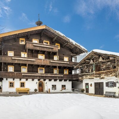 The traditional Farm House, featuring dark wooden facades and balconies, stands next to a rustic outbuilding, all surrounded by snow-covered hills.