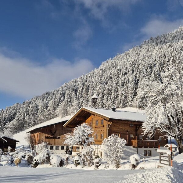 The farmhouse in winter, surrounded by snow-covered trees and mountains.