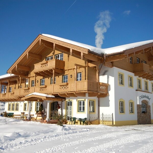The Farm House exterior, showcasing traditional wooden balconies and a snow-covered landscape under a clear blue sky, with smoke rising from the chimney.
