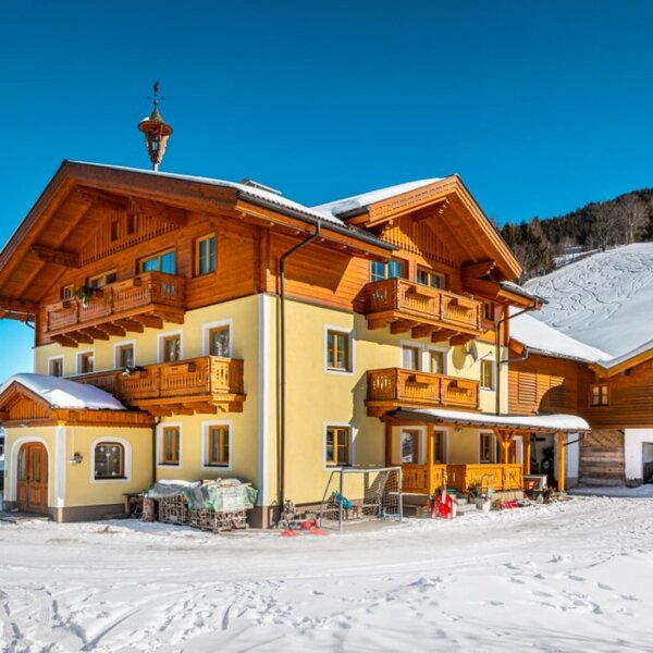 The farm house features a light yellow facade with multiple wooden balconies, situated in a snowy mountain landscape.