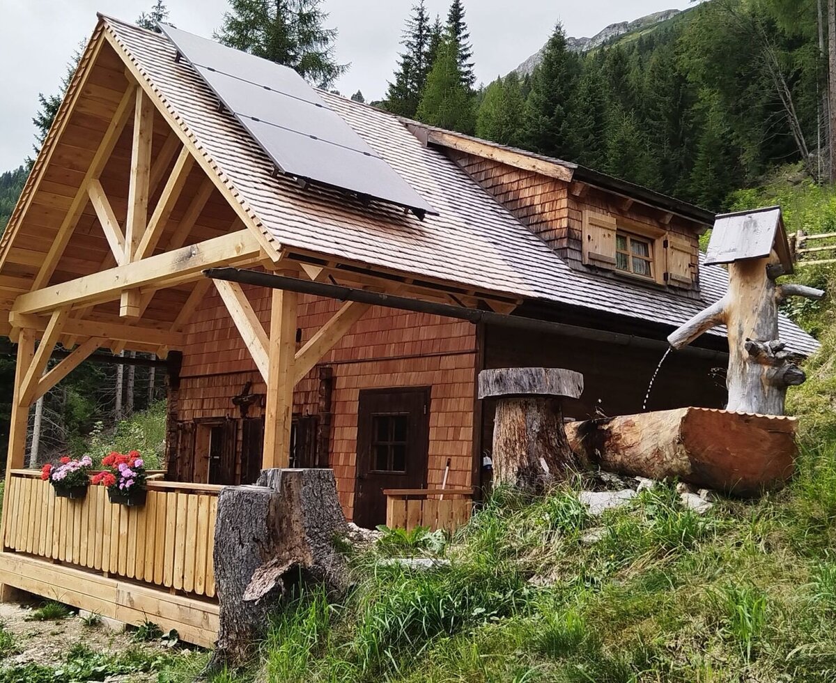 The wooden farmhouse features a covered terrace, solar panels on the roof, and a rustic log fountain.
