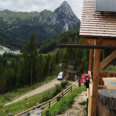 The farmhouse features solar panels on its roof and a covered terrace, accessible by a dirt road, offering views of the mountain, forest, and Lake Schlierersee.