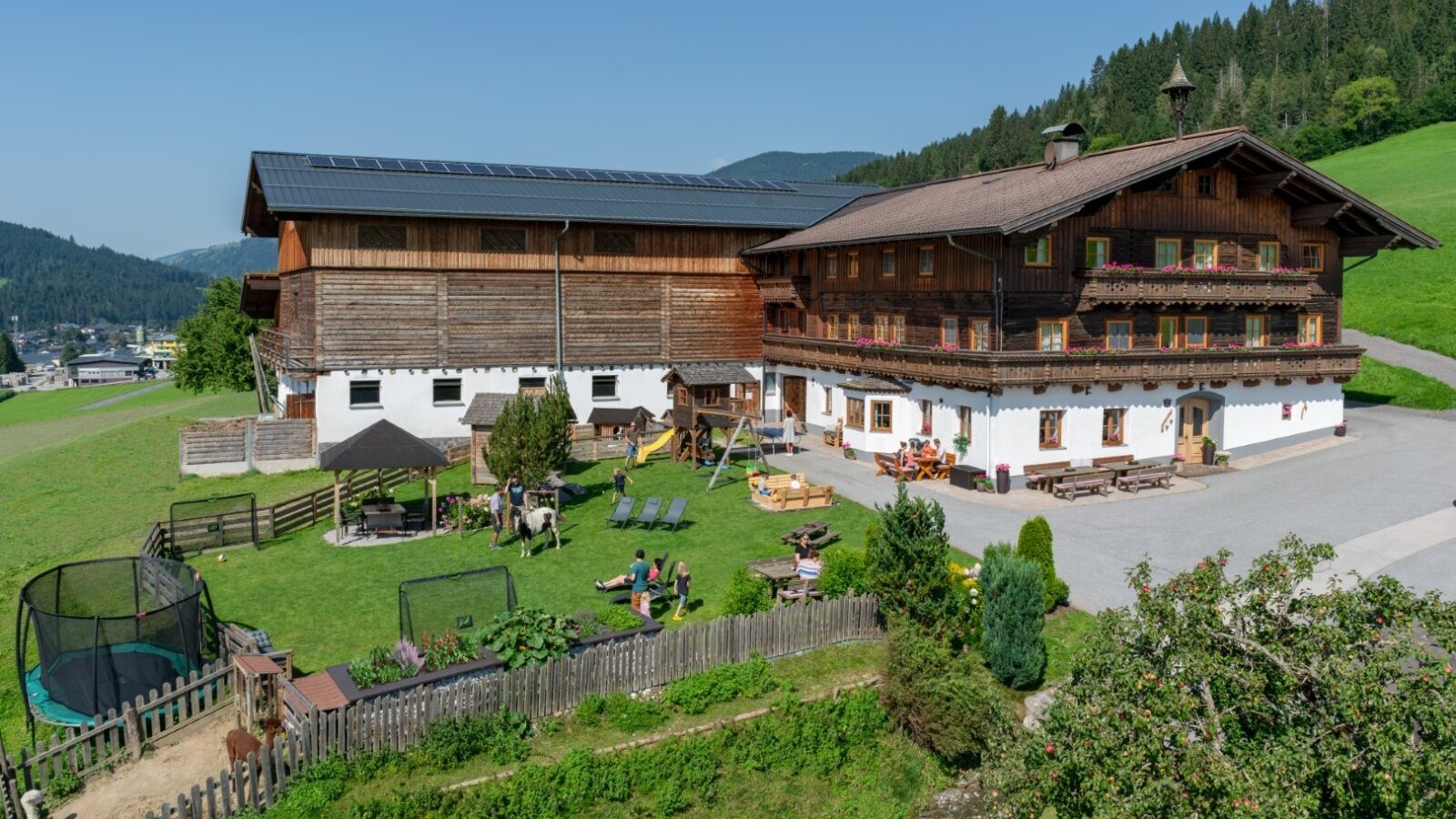 Exterior view of the farmhouse featuring a large garden, children's playground, trampoline, covered seating area, and animal enclosures.