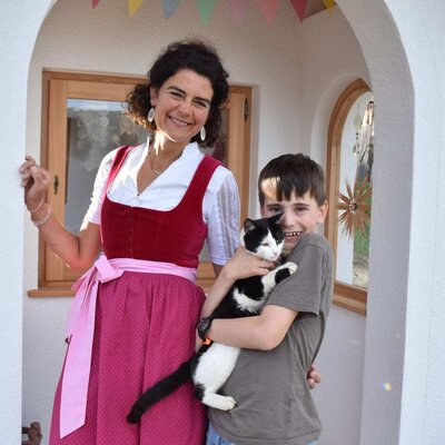 The host family of the farmhouse welcomes guests, featuring a woman in traditional dress and a child with a cat.