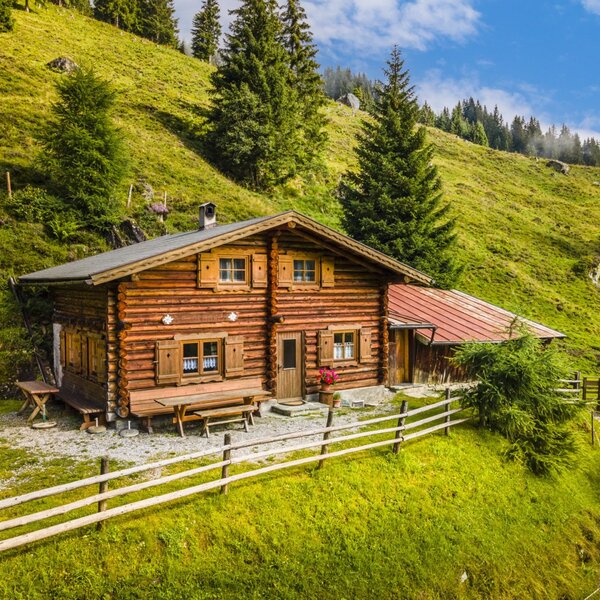 The log-built farmhouse includes wooden shutters and an outdoor picnic table, surrounded by a wooden fence and green hills.