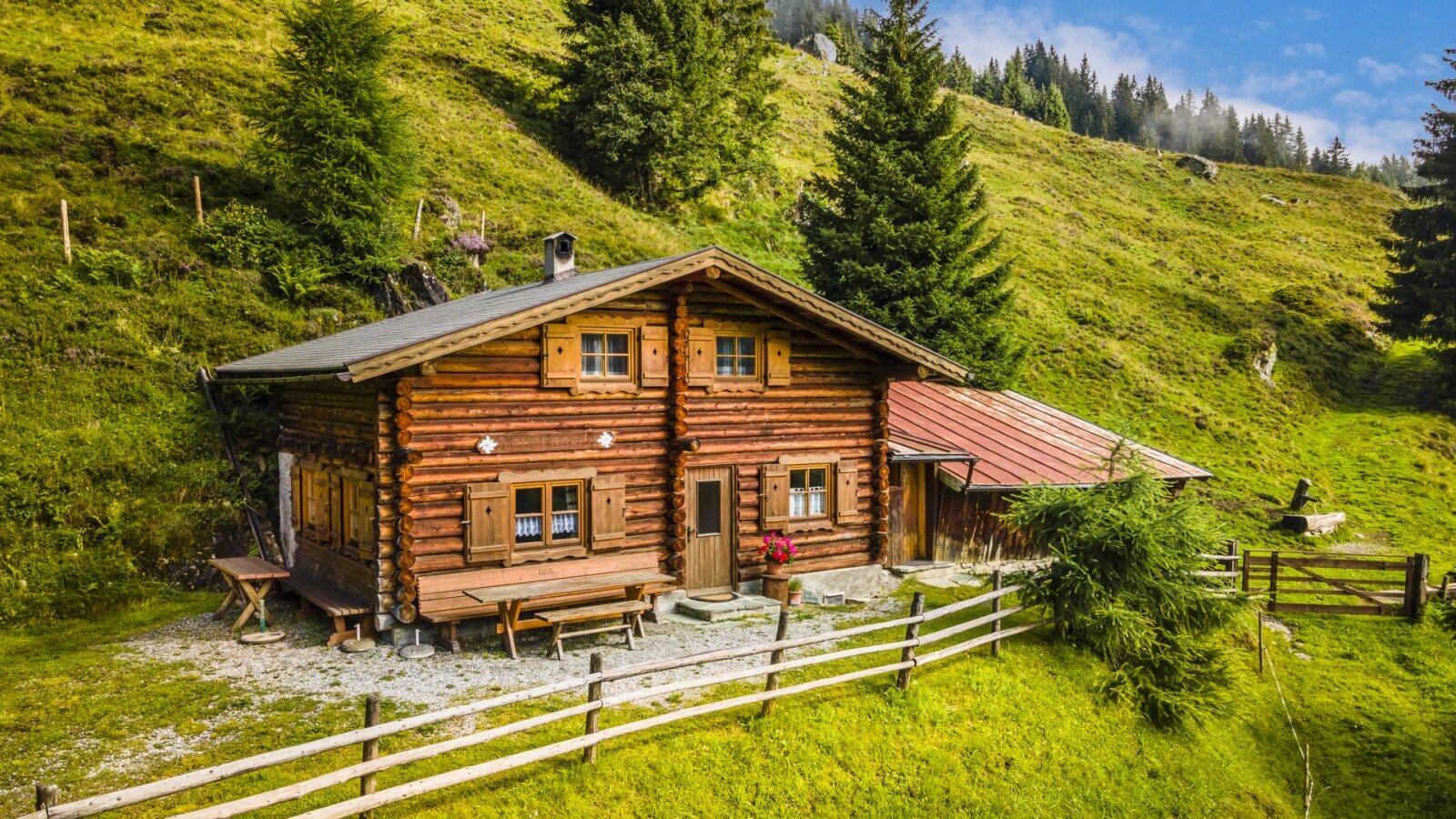 The log-built farmhouse includes wooden shutters and an outdoor picnic table, surrounded by a wooden fence and green hills.