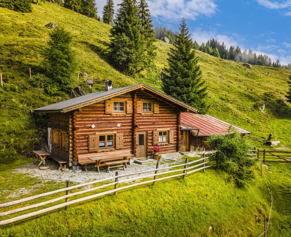 The log-built farmhouse includes wooden shutters and an outdoor picnic table, surrounded by a wooden fence and green hills.