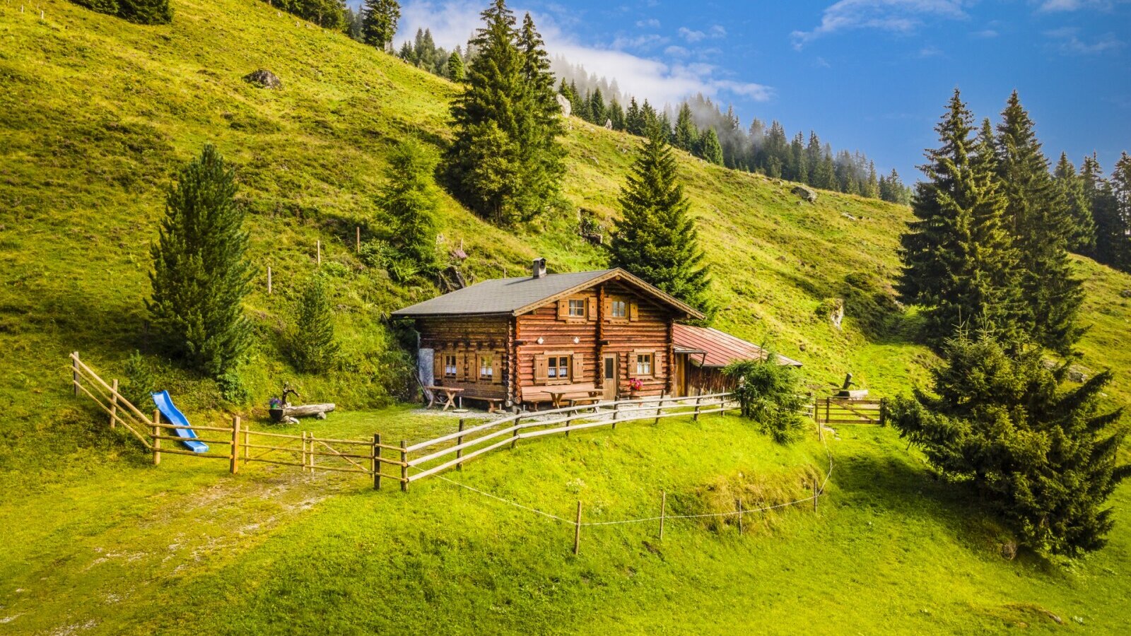 The farmhouse exterior, a log cabin structure on a grassy hillside, surrounded by pine trees and featuring a children's slide.