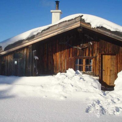 The wooden exterior of the farmhouse in winter, with snow covering the roof and ground, and a path leading to the front door.