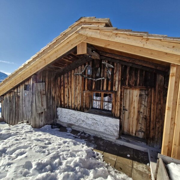 Exterior view of the Hut with a wooden facade, wooden door, and snowy surroundings against a mountain backdrop.