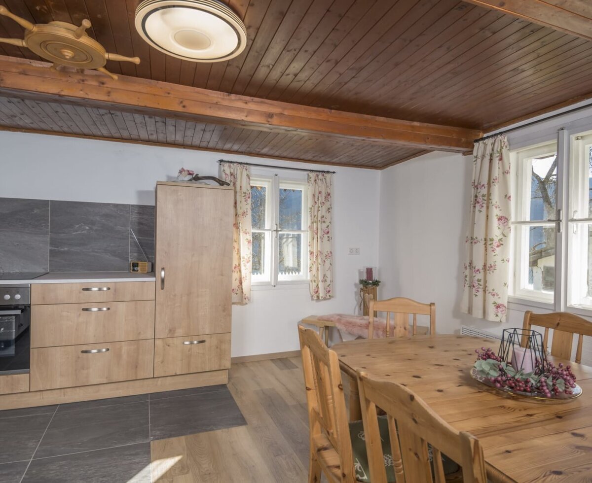 A cozy kitchen-cum-living room in the Alm, featuring a bright wooden dining area, rustic timber ceiling, and a modern kitchenette.