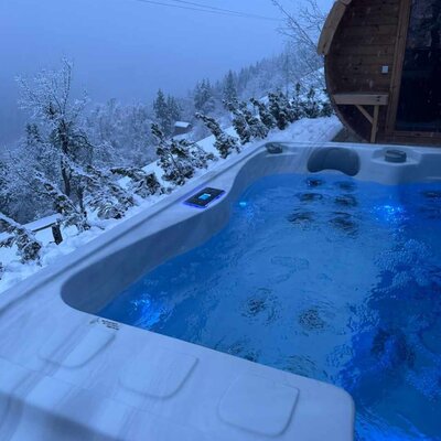 The Alm's inviting outdoor hot tub with illuminated blue water, set against a snowy winter landscape next to a cozy barrel sauna.