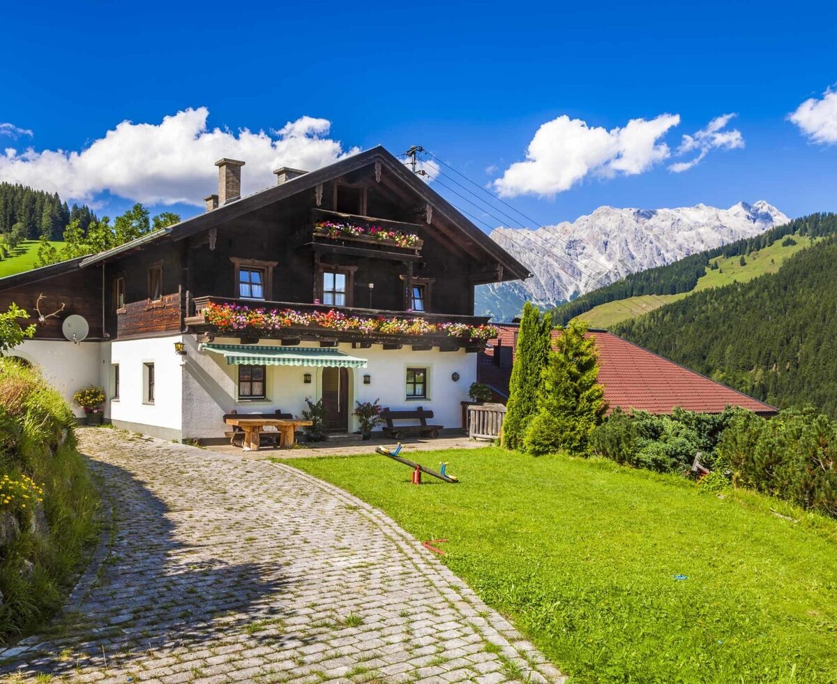 The farm house exterior features flower-filled balconies, a paved entrance path, a garden with a seesaw and outdoor seating, all set against a mountain backdrop.
