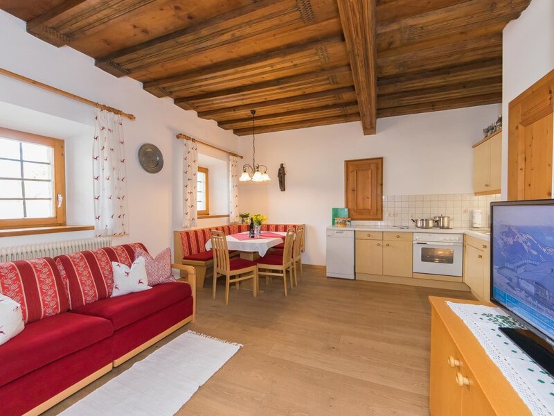 Living area in an apartment of the Farm House, featuring a red sofa, a dining corner, a fully equipped kitchen, and a television, all beneath wooden ceiling beams.