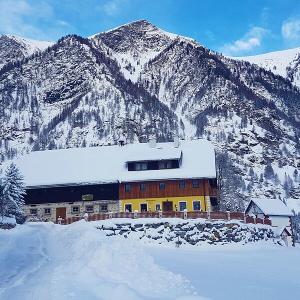 The farm house exterior, covered in snow, set against a backdrop of towering, snow-capped mountains.