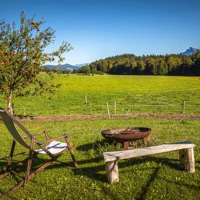 An inviting relaxation spot at the farmhouse with a wooden deck chair, a rustic bench, and a fire pit overlooking a vast green meadow.