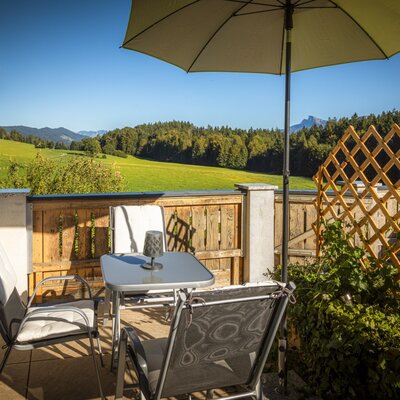 The farmhouse's sunny balcony features cozy outdoor seating, a large parasol, and wide views of the green meadows and forests.