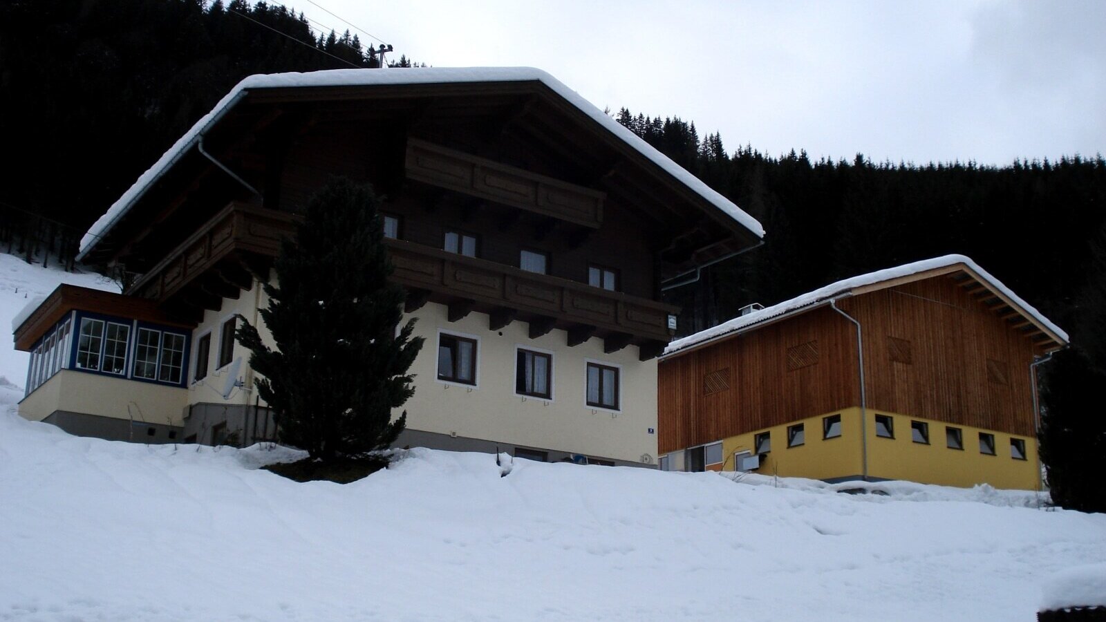 The farm house exterior, featuring traditional wooden balconies, a glass-enclosed extension, and an adjacent wooden and yellow building, all set in a snow-covered landscape.