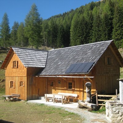 Exterior of the farmhouse, featuring a wooden facade, shingle roof with a solar panel, and an outdoor seating area with benches, set against a forest.