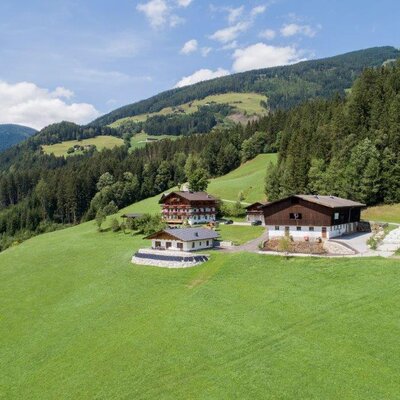 The farmhouse with its buildings, surrounded by green meadows, forested mountains, and hay bales.