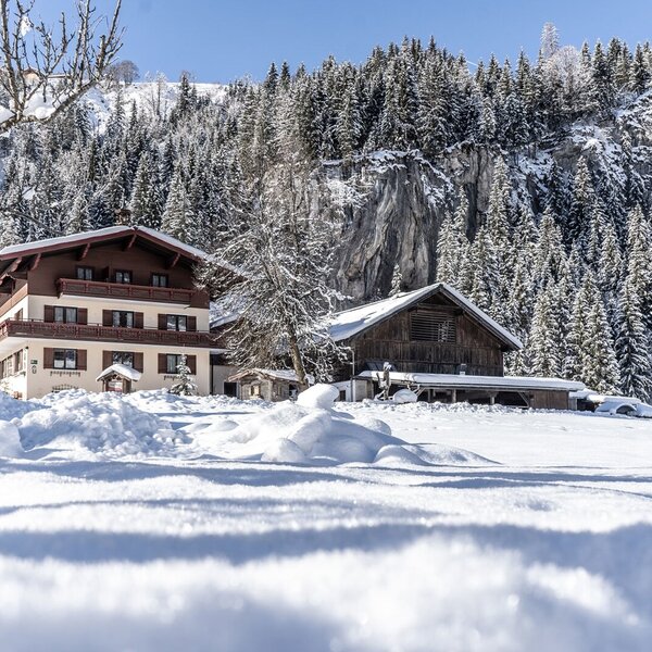 The farm house, featuring a multi-story main building with balconies and an adjacent wooden barn, is set amidst snow-covered mountains and evergreen forests.