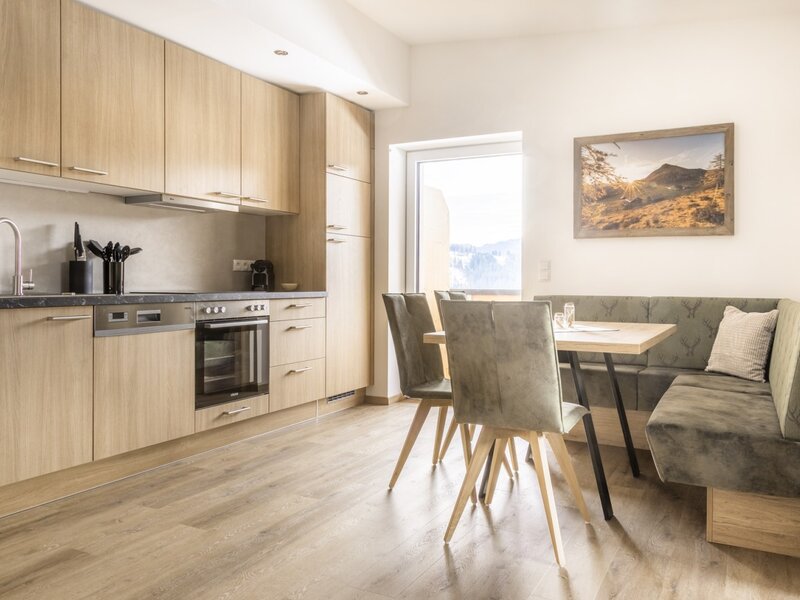 Modern kitchen with wooden elements, oven, dishwasher, and dining area with corner bench and outdoor view.