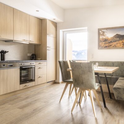 Modern kitchen with wooden elements, oven, dishwasher, and dining area with corner bench and outdoor view.