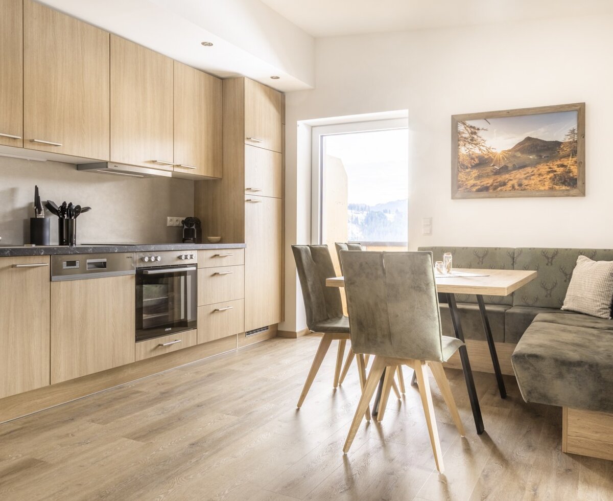 Modern kitchen with wooden elements, oven, dishwasher, and dining area with corner bench and outdoor view.