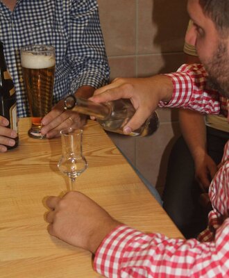 A guest pouring a clear beverage into a small glass at a wooden table, with another guest holding a beer in the Farm House.