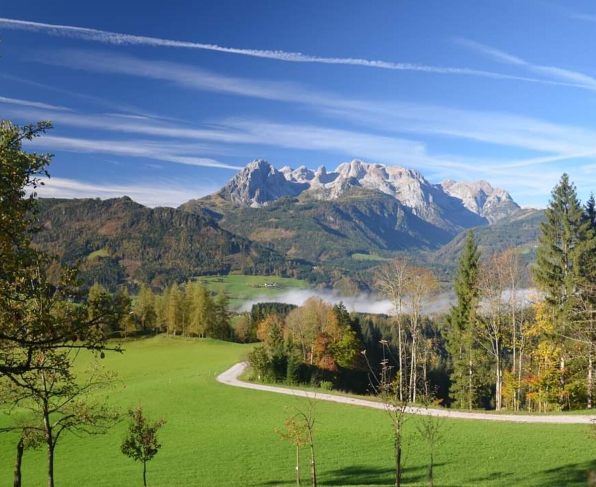 The mountainous landscape surrounding the farmhouse, featuring rocky peaks, green fields, autumn foliage, and a road winding through the valley mist.