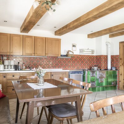 Kitchen and dining area of the farmhouse, featuring wooden beams, a tiled kitchen counter, a green tiled stove, and a dining table with bench seating.