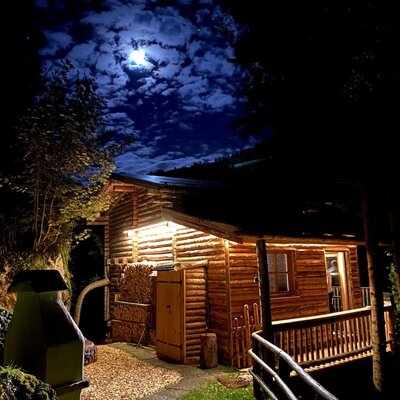 An inviting Alm at night, warmly lit under a dramatic moonlit sky.