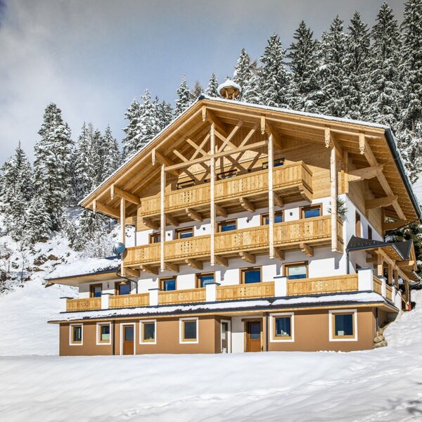 The farmhouse exterior, featuring multiple wooden balconies and a distinct gabled roof, nestled among snow-covered pine trees.