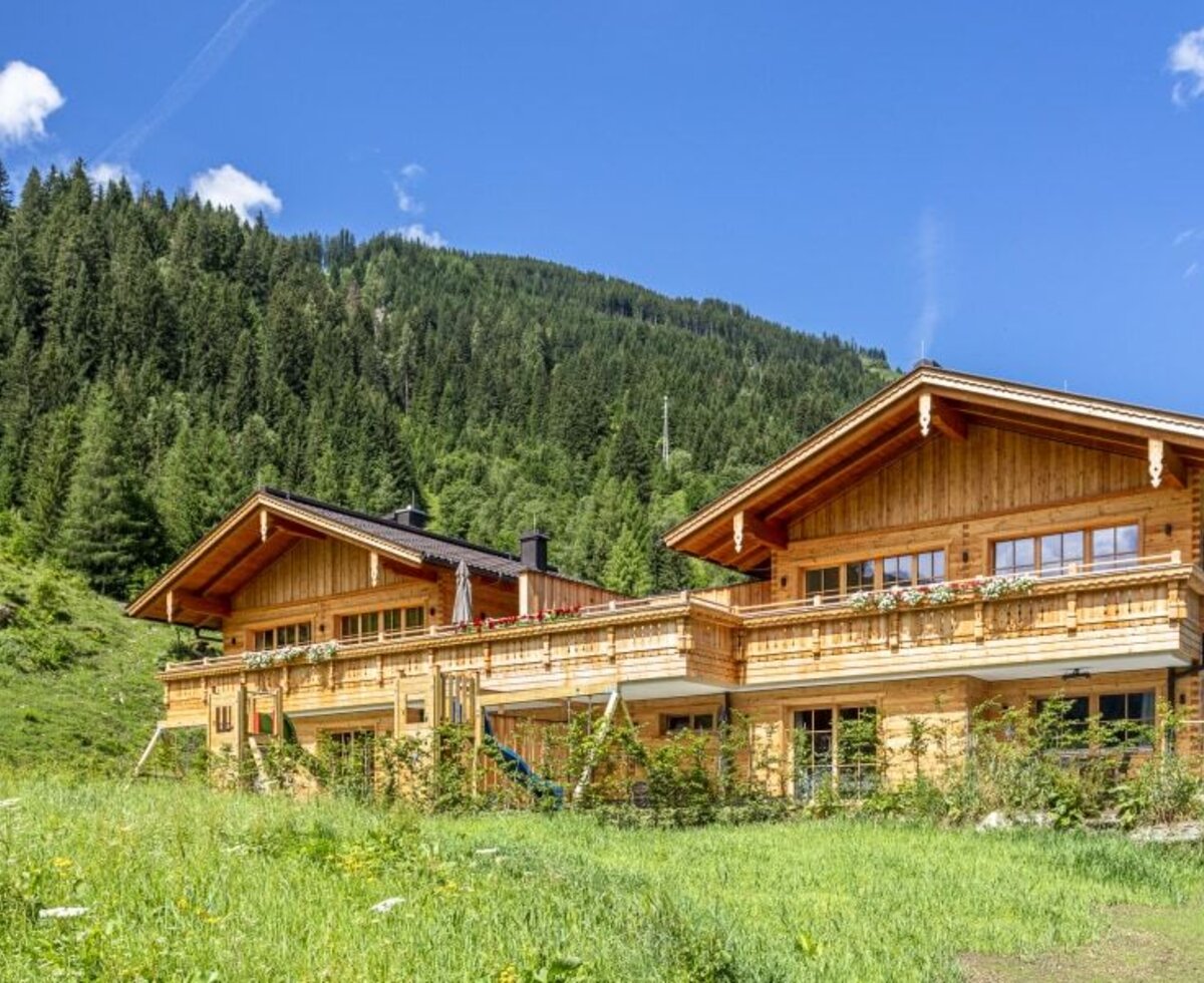 The wooden farmhouse, featuring balconies with flower boxes, is located on a green hillside surrounded by forests, with a paved road leading to the property.