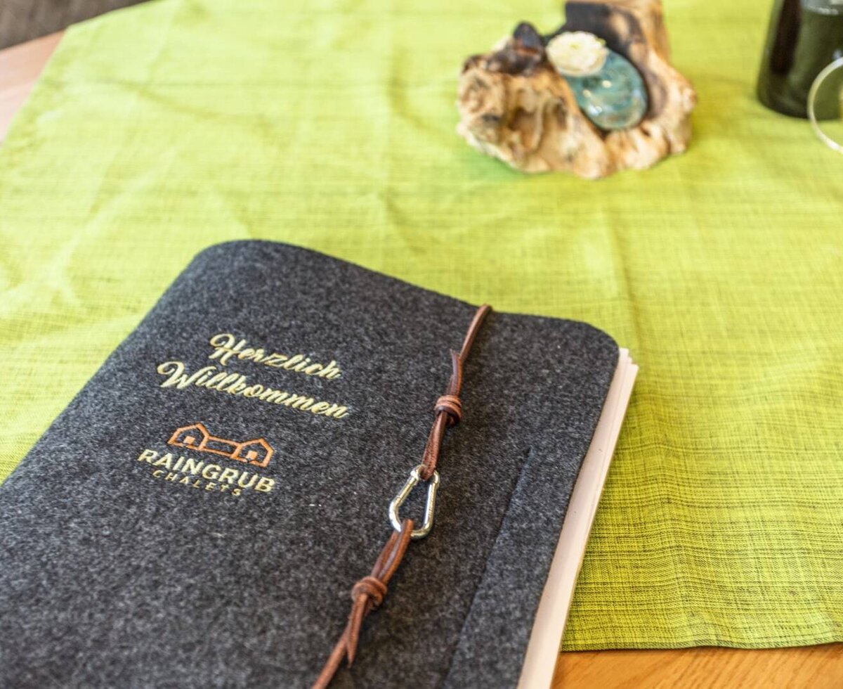 The welcome folder for the farmhouse, featuring a grey felt cover with 'Herzlich Willkommen' and 'RAINGRUB CHALETS' branding.