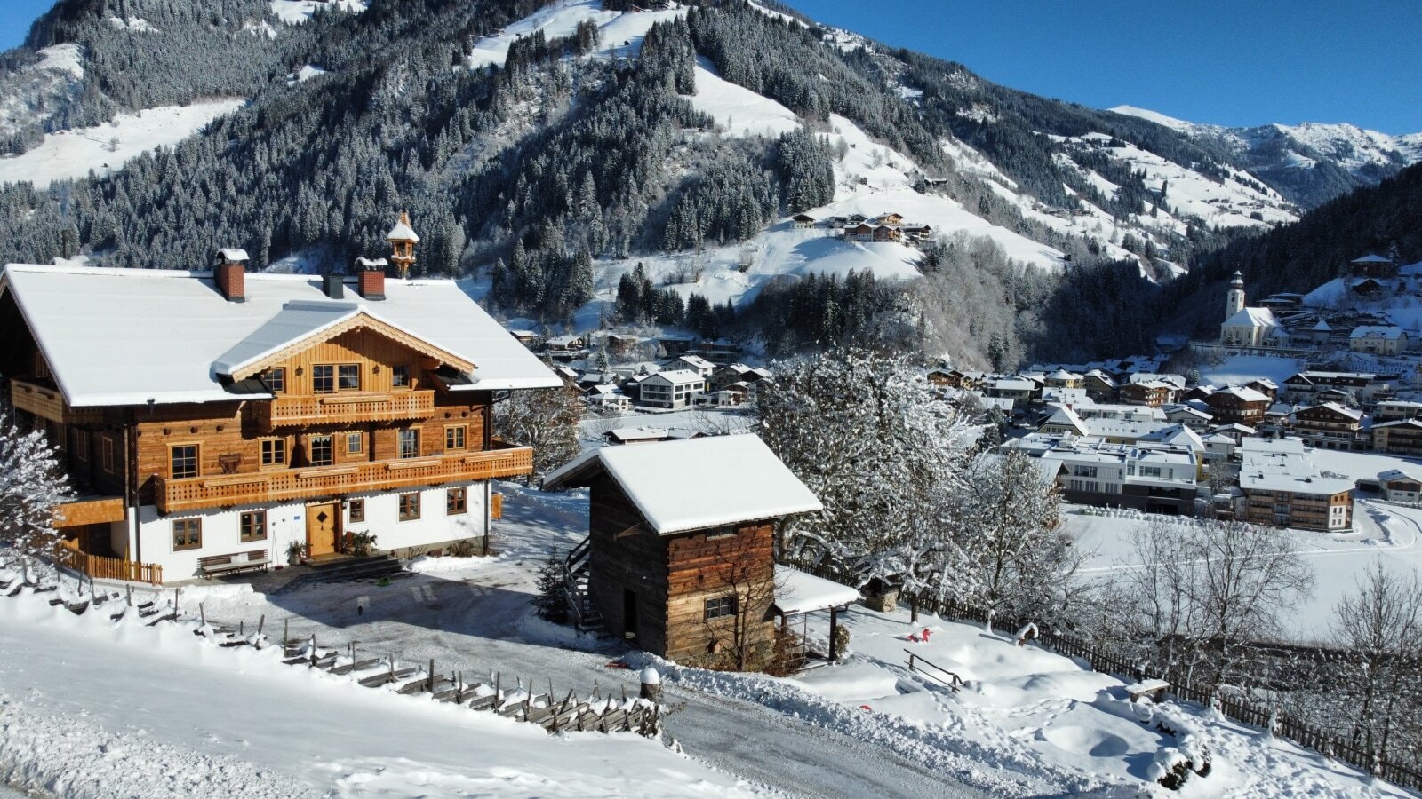 The farmhouse with wooden balconies in winter, surrounded by snow and with a view of the valley and mountains.