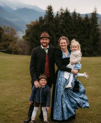 The host family of the farmhouse in traditional attire with children, set against a mountain backdrop.
