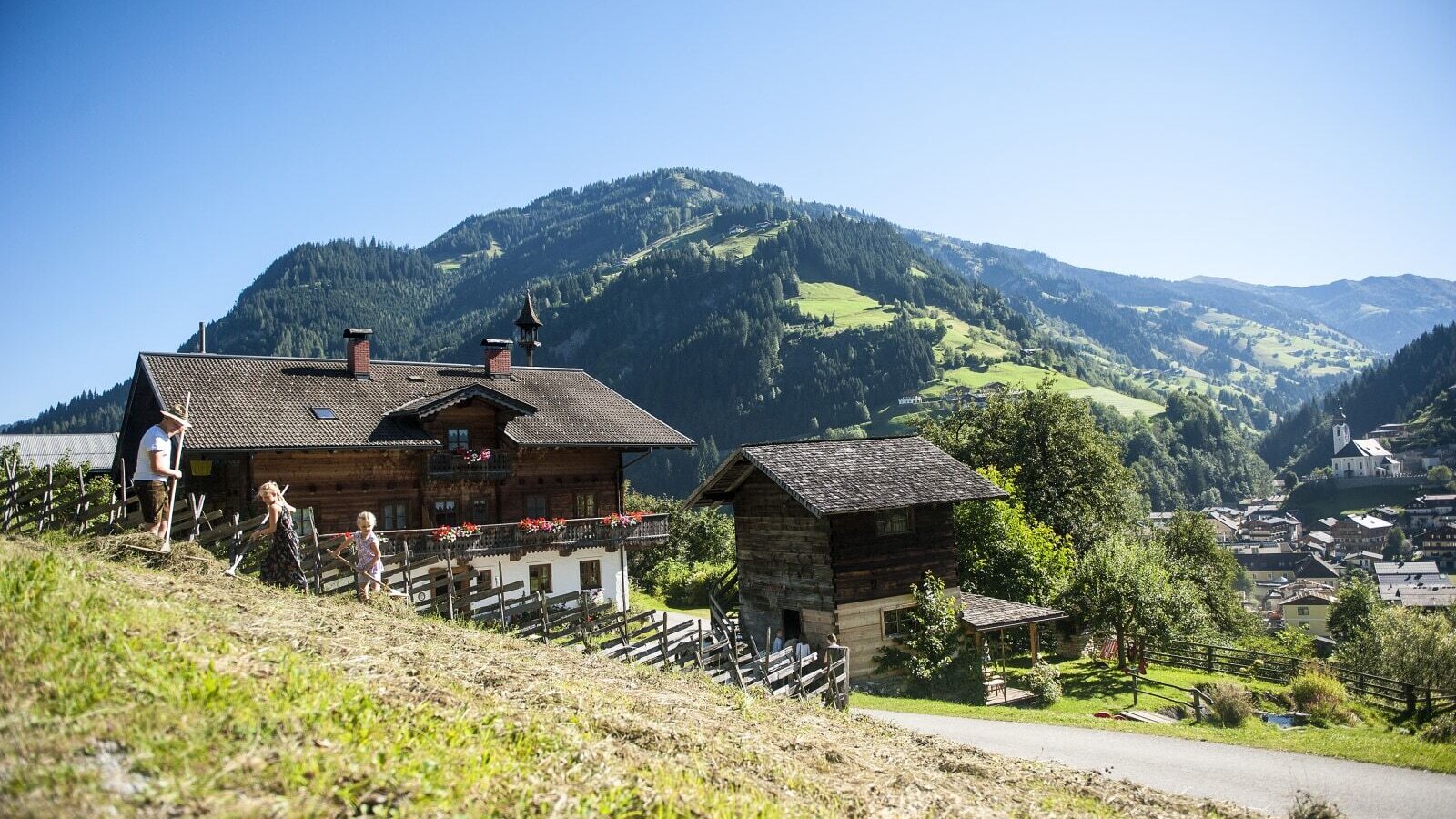 The traditional wooden Farm House is set against a backdrop of green mountains and a distant village, showing people engaged in outdoor farm activities.