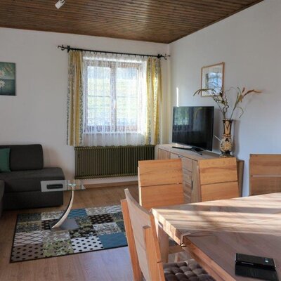 The living room of the farmhouse includes a corner sofa, a dining area with a wooden table, and a television.