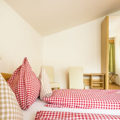 Guest bedroom in the farm house with a bed, red and beige checkered bedding, a wooden wardrobe featuring a mirrored panel, and a small desk with two chairs.