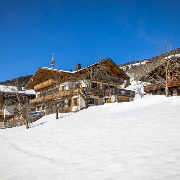 The exterior of the farm house, with its wooden balconies and weathervane, is set against a snow-covered hillside with distant mountains.