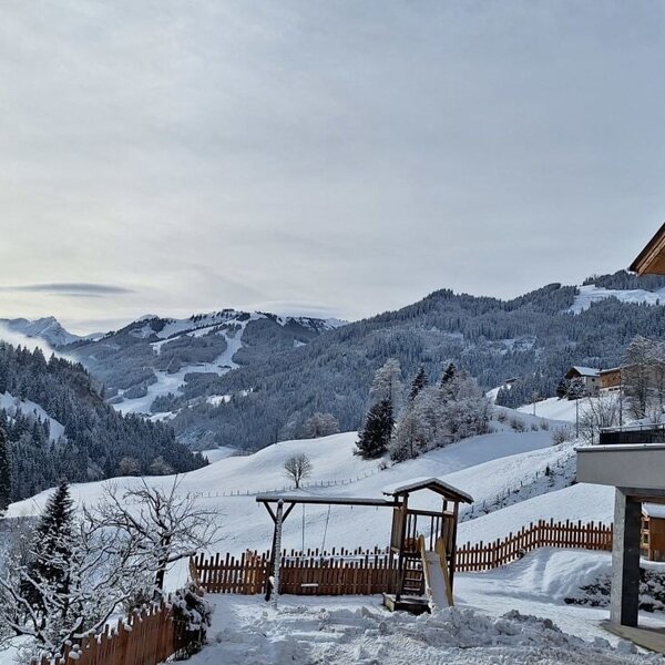 The exterior of the farm house featuring wooden balconies, a snow-covered children's playground, and views of the surrounding snowy mountains.